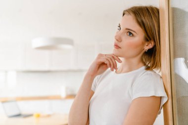 Evdeki mutfakta duvara yaslanan düşünceli genç bir kadın.