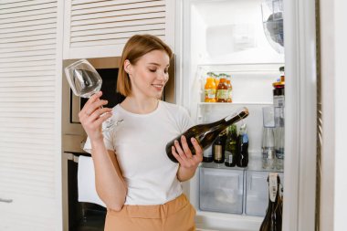 Gülümseyen sarışın beyaz kadın. Günlük kıyafetler içinde. Elinde şampanya şişesi ve bardakla evdeki açık buzdolabının önünde duruyor.