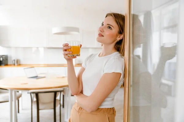 微笑着的年轻女人倚在厨房的墙上 手里拿着一杯果汁 — 图库照片