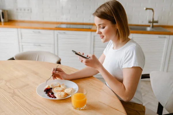 坐在厨房里 拿着手机 笑着吃着健康早餐的年轻女人 — 图库照片