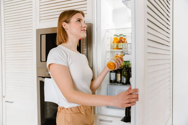 年轻的金发女人站在冰箱边 手里拿着一瓶天然果汁 — 图库照片