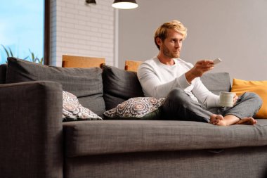 Sarışın adam evde kanepede otururken çay içip televizyon seyrediyor.