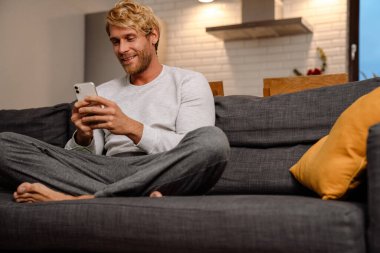 Sarışın sakallı adam evdeki koltukta otururken cep telefonu kullanıyor.