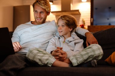 Kızıl adam oğluyla evde kanepede dinlenirken dizüstü bilgisayar kullanıyor.