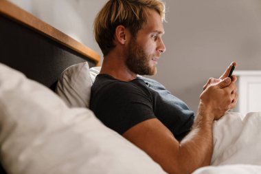 Sarışın sakallı adam evde yatarken cep telefonu kullanıyor.
