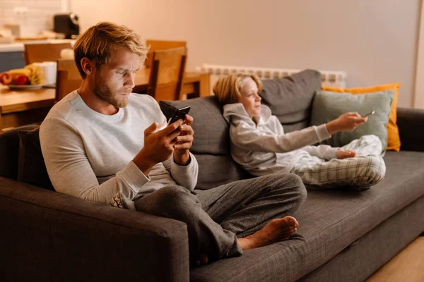 白父母坐在沙发上 一边用手机 一边看电视 — 图库照片