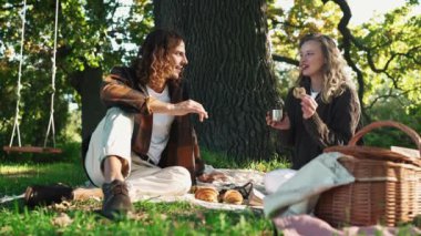 Neşeli genç çift sıcak çay içiyor ve parkta piknikte konuşuyor.