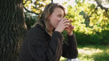 Gülümseyen sarışın kadın çay içiyor ve parkta piknikte kameraya bakıyor.