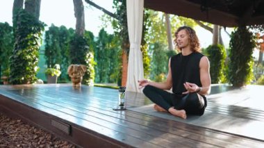 Kıvırcık saçlı rahat sporcu dışarıda yoga minderinde lotus pozisyonunda meditasyon yapıyor.