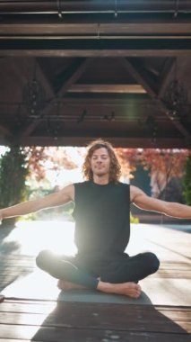 Kıvırcık saçlı güzel atletik adam dışarıda yoga minderinde lotus pozisyonunda meditasyon yapıyor ve nefes alıyor.