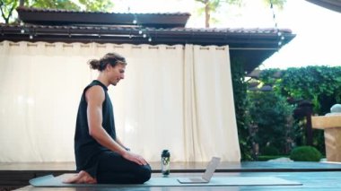 Gülümseyen atletik kıvırcık saçlı adam bilgisayarla konuşuyor ve yoga minderinde meditasyon yapıyor.