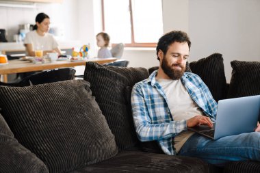 Orta yaşlı bir adam dizüstü bilgisayarda çalışıyor. Evdeki koltukta oturuyor. Karısı ve küçük oğlu arka plandaki mutfak masasında kahvaltı yapıyor.