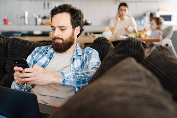 Middle aged brearded man working on laptop computer holding mobile phone sitting on a couch at home his wife and little son having breakfast at the kitchen table on a background