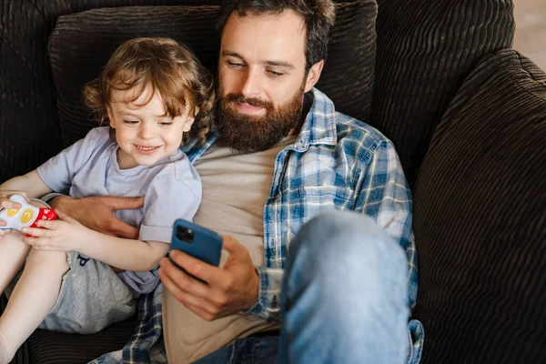 在家里和小儿子坐在沙发上 一边笑一边看手机上的视频 — 图库照片