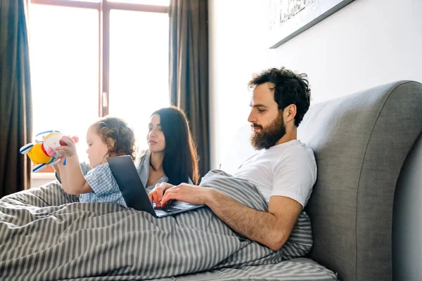 爸爸在笔记本电脑上工作 快乐的一家人一起在床上放松 — 图库照片