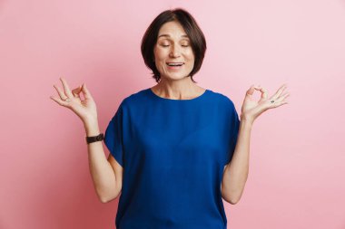 Olgun esmer kadın gülümserken pembe arka planda zen hareketi yapıyor.