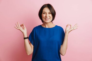 Olgun esmer kadın gülümserken pembe arka planda zen hareketi yapıyor.