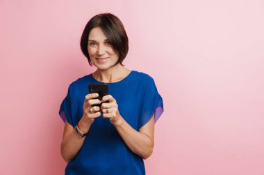 Pembe arka planda izole edilmiş gülümseyen ve cep telefonu kullanan olgun esmer kadın.