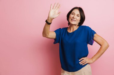 Olgun esmer kadın pembe arka planda izole edilmiş kameraya el sallarken gülüyor.