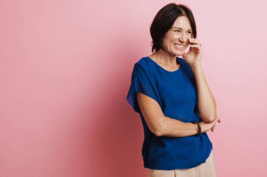 Olgun esmer kadın, pembe arka planda izole edilmiş yüzünü kapatırken gülüyor.