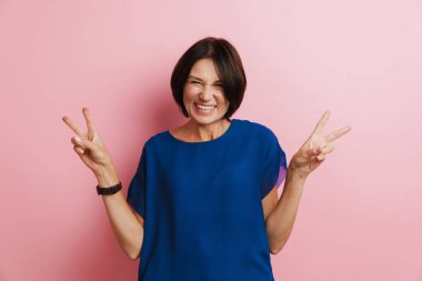 Olgun bir kadın pembe arka planda yalnız başına kameraya gülerken barış jesti yapıyor.