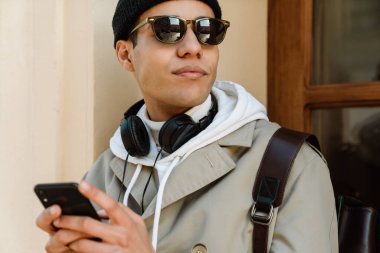 Dışarıda duvarın yanında duran güneş gözlüğü takan genç adam cep telefonu kullanıyor.