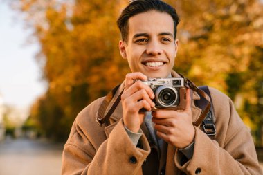 Paltolu genç adam sonbahar parkında yürürken retro kamerada fotoğraf çekiyor.
