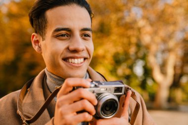 Paltolu genç adam sonbahar parkında yürürken retro kamerada fotoğraf çekiyor.