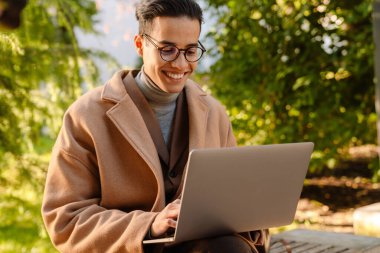 Paltolu genç adam dışarıda parkta bankta otururken dizüstü bilgisayarla çalışıyor.