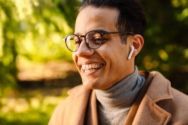 Genç adam gözlük takıyor müzik dinliyor ve parkta gülüyor.