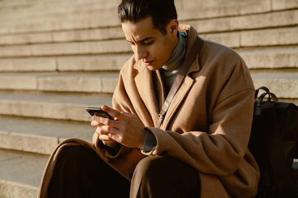 Молодий Чоловік Пальто Використовуючи Мобільний Телефон Сидячи Сходах Відкритому Повітрі — стокове фото