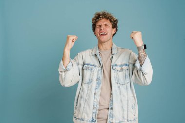 Ceket giyen genç kıvırcık adam çığlık atarak mavi arka plandan izole edilmiş bir jest yapıyor.