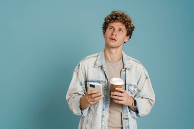 Genç kıvırcık adam mavi arka planda izole kahve içerken cep telefonu kullanıyor.