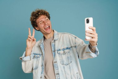Genç adam mavi arka planda cep telefonuyla selfie çekerken barış jesti yapıyor.