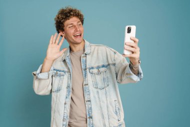 Genç kıvırcık adam mavi arka planda izole edilmiş cep telefonuyla selfie çekerken el sallıyor.