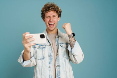 Genç kıvırcık adam mavi arka planda izole bir şekilde cep telefonuyla oyun oynarken kazanan hareketi yapıyor.