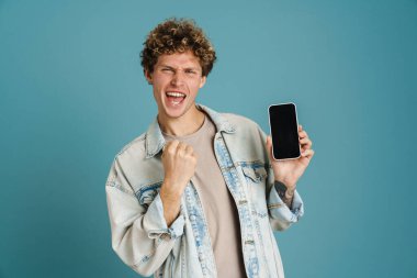 Genç kıvırcık adam cep telefonunu gösteriyor ve mavi arka planda izole edilmiş bir jest yapıyor.