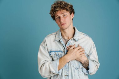Mavi arka planda, elleri göğsünde poz veren kıvırcık ceketli genç bir adam.