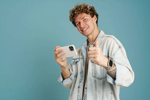 年轻的卷发男子在使用蓝色背景的手机时露出大拇指 — 图库照片