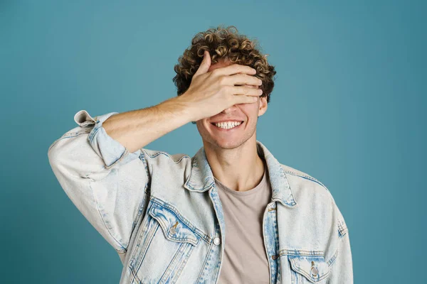 Young man wearing jacket smiling while covering his eyes isolated over blue background
