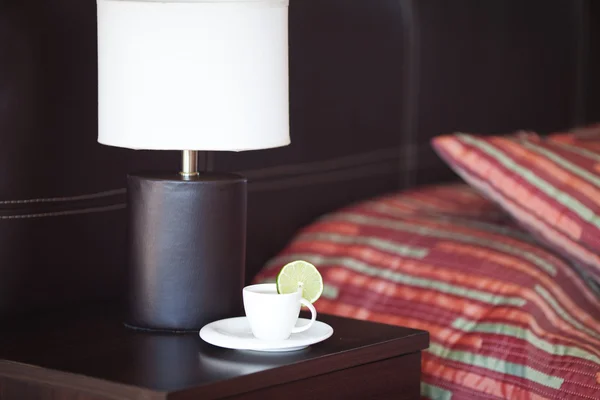 Cama com um travesseiro, uma xícara de chá na mesa de cabeceira e lâmpada — Fotografia de Stock