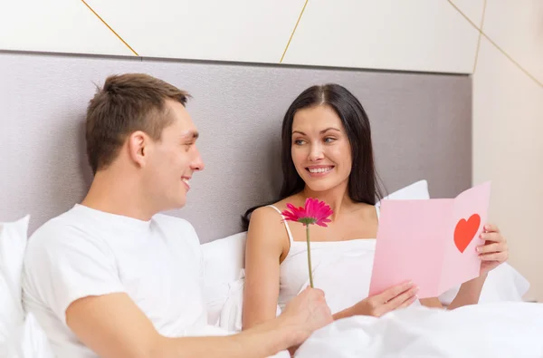 Casal sorridente na cama com cartão postal e flor — Fotografia de Stock