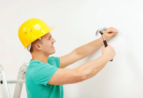 Hombre sonriente en casco clavo de martilleo en la pared — Foto de Stock