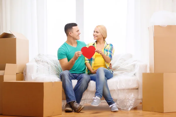 Pareja sonriente con el corazón rojo en el sofá en el nuevo hogar — Foto de Stock