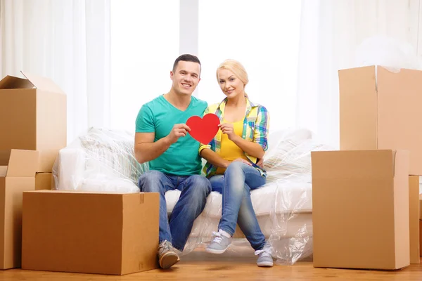 Pareja sonriente con el corazón rojo en el sofá en el nuevo hogar — Foto de Stock