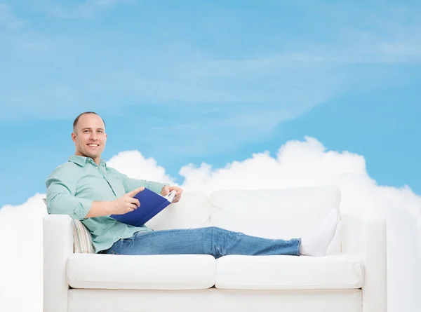 Homme souriant couché sur un canapé avec un livre — Photo