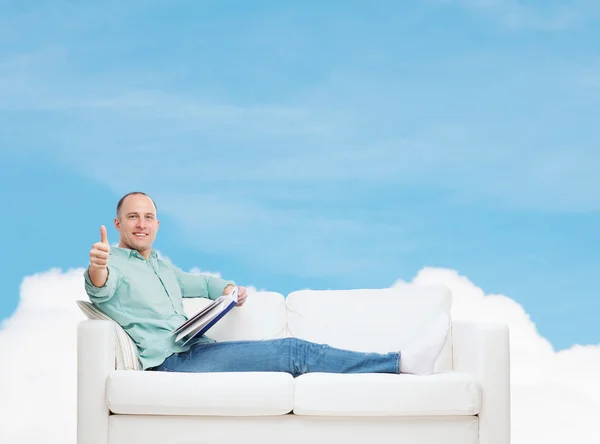 Homme souriant couché sur un canapé avec un livre — Photo