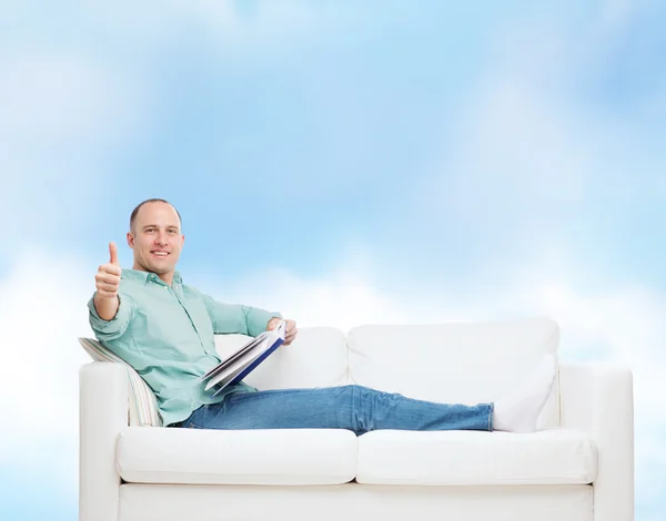 Homme souriant couché sur un canapé avec un livre — Photo