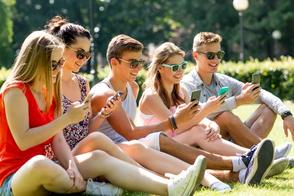Улыбающиеся друзья со смартфонами, сидящими на траве — стоковое фото