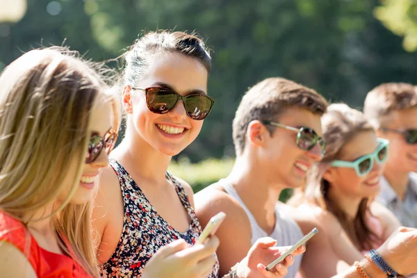 Amigos sonrientes con teléfonos inteligentes sentados en el parque — Foto de Stock